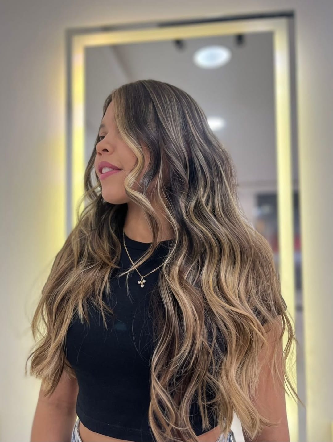 Cabelo castanho longo com mechas iluminadas e ondas, diante do espelho com luz dourada do salão
